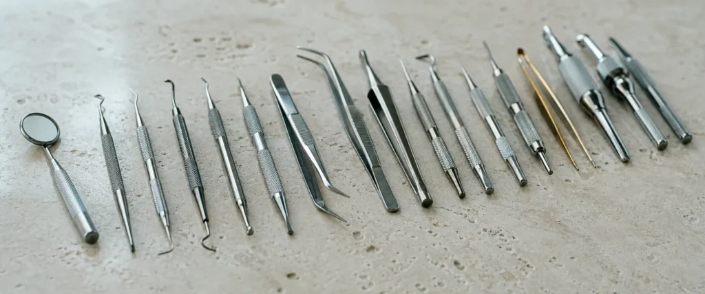 A long row of polished stainless-steel dental instruments on pale limestone